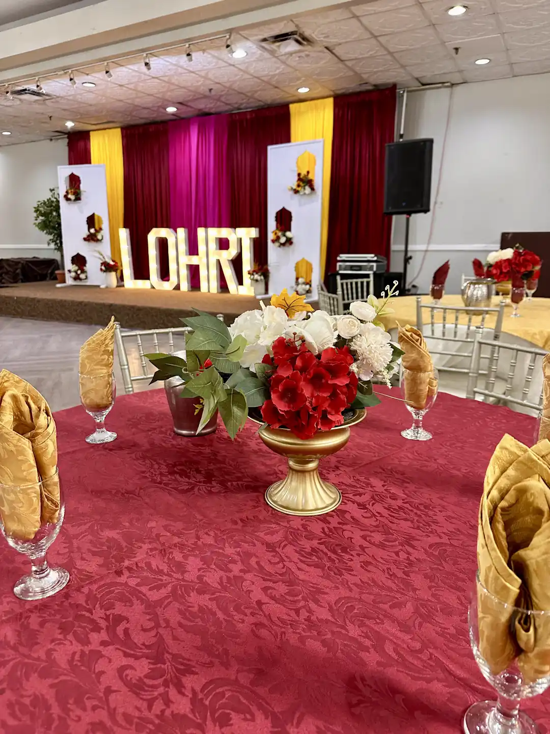 Lohri celebration hall with red damask tablecloth, gold floral centrepiece, and red and gold stage backdrop with marquee letters