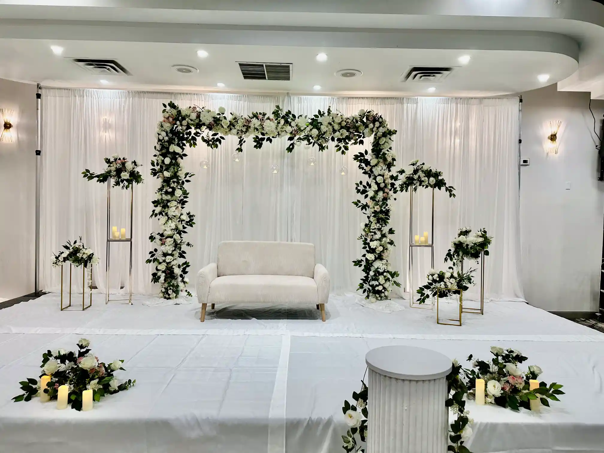 White fabric backdrop with large white floral arch, cream loveseat, and gold floral stands for a wedding stage